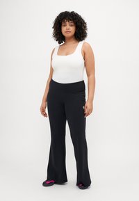 Fehér négyzet alakú nyakú bodysuit fekete, bővülő nadrággal párosítva, melynek rugalmas derékrésze és logó részlete van. A modellen fekete papucsok láthatók, rózsaszín kiegészítőkkel.