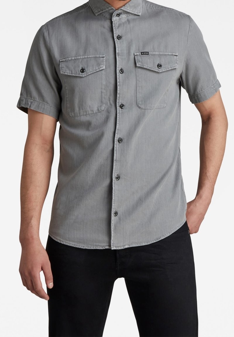 Homme portant une chemise grise à manches courtes avec deux poches poitrine boutonnées et un pantalon noir, debout sur un fond blanc.