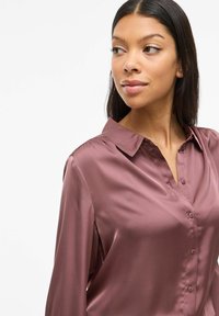 Camicia in satin mauve con colletto, maniche lunghe e chiusura con bottoni. Tessuto liscio, caratterizzato da un leggero lucido.