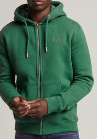 Felpa verde con zip, dotata di cordini, tasche a marsupio e un accento con logo. Realizzata in morbido tessuto texturizzato, presenta polsini e orlo a costine.