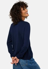 Pull-over long bleu marine à manches longues avec un col rond et des poignets côtelés, associé à un jean bleu clair, vu de dos avec les mains croisées.