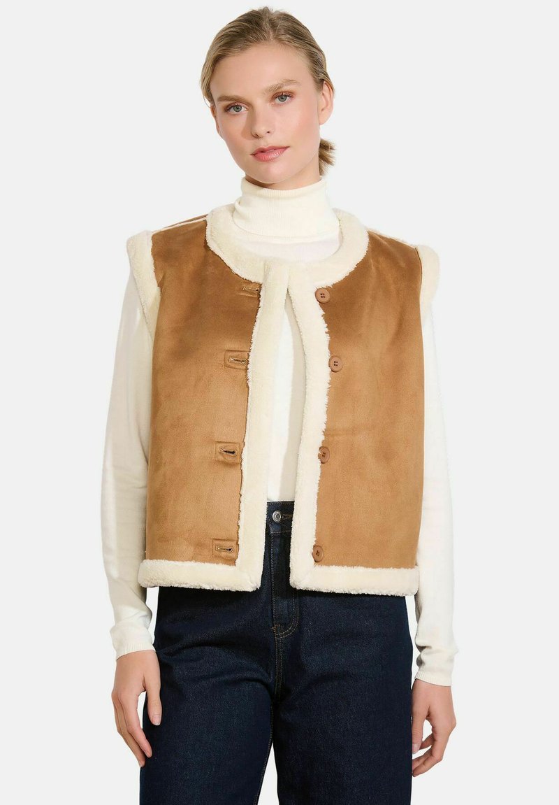 Ženska nosi svetlo rjav suede jopič z belim ovitkom iz ovčje kože čez belo turtleneck majico in temne kavbojke, stoji ob navadni ozadju.