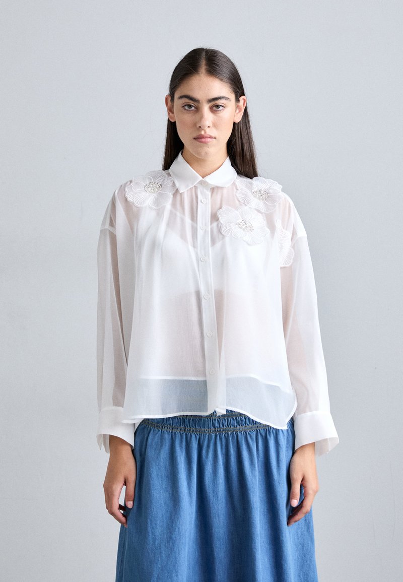 Hvid, gennemsigtig skjorte med knapper og broderede blomstermotiver på skuldrene, parret med en blå denimnederdel med elastisk talje.