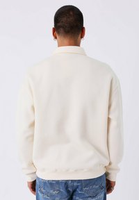 Beige oversized sweatshirt met een zachte textuur en kraag. Beschikt over geribde boorden en zoom, gedragen met lichtblauwe denim met een logo-patroon.