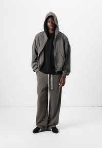 Grauer oversized Zip-Hoodie mit Kapuze, schwarzes T-Shirt darunter, graue Jogginghose mit seitlichen Knöpfen und schwarze Slipper.