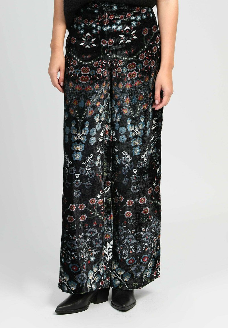 Pantalons en velours noir à jambes larges avec des motifs floraux colorés, portés avec des bottes à talons noirs à bout pointu, vue partielle de la personne de la taille aux pieds.