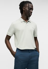 Ανοιχτοπράσινο πόλο με κοντά μανίκια, κλασικό γιακά και διακριτικό λογότυπο στο στήθος, συνδυασμένο με σκούρο μπλε παντελόνι, παρουσιάζοντας άνετη εφαρμογή.