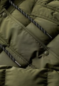Olive green puffer jacket close-up with quilted texture, features black drawcord with white accents, and a round button detail.