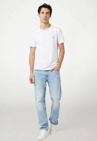 T-shirt bianca a maniche corte con piccolo logo rosso sul petto, abbinata a jeans blu chiaro a gamba dritta e sneaker bianche.