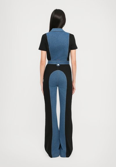 Žena s dlouhými tmavými vlasy stojí čelem k nám, má na sobě široké kalhoty z černého a modrého denimu ve stylu colorblock a odpovídající zkrácenou vestu přes černé tričko.