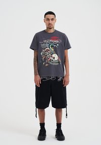 Harmaa t-paita, jossa on monivärinen tatuointityylinen printti ja teksti "Ed Hardy". Yhdistetty mustiin cargo-shorteihin ja mustiin lenkkareihin.