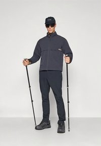 Granatowa kurtka na zamek z kontrastową panelową częścią na piersi, w połączeniu z ciemnymi spodniami i butami do wędrówek. Postać trzyma kijki trekkingowe i nosi czapkę oraz okulary przeciwsłoneczne.