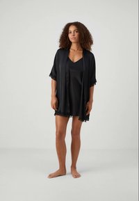 Femme aux cheveux bouclés, pieds nus, portant une robe en satin noire avec une robe de chambre de style kimono assortie, contre un fond uni.