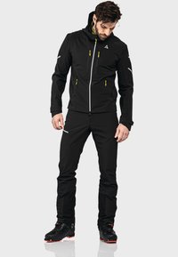 Schwarze wasserdichte Jacke mit gelben Reißverschlüssen und reflektierenden Akzenten, kombiniert mit schwarzen Hosen und schwarzen Stiefeln mit roten Sohlen, entworfen für Outdoor-Aktivitäten.