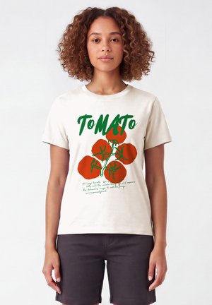 Femme aux cheveux bouclés portant un T-shirt blanc avec un graphisme de tomate verte et rouge et le mot "Tomato" en grandes lettres.