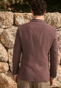Blazer marron à texture avec une coupe ajustée, fente simple au dos et tissu contrastant aux poignets, associé à un pantalon clair.