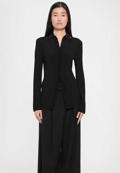 Cardigan nero a coste con chiusura a bottoni, maniche lunghe e colletto a punta. Presenta uno spacco laterale e una texture liscia.