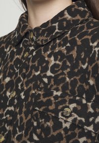 Chemise en imprimé léopard avec une palette de couleurs noire et brune, réalisée en tissu doux avec une texture légèrement duveteuse et des accents de boutons.