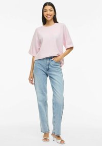 Ohlapno svetlo roza pleteno pulover z kratkimi rokavi, kombiniran z visokim pasom svetlo modrih kavbojk z ravnimi hlačnicami in belimi sandali s peto.