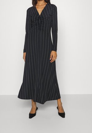 Kobieta ubrana w czarną sukienkę midi z długim rękawem, ozdobioną pionowymi białymi cienkimi paskami i marszczeniem na wysokości piersi, w zestawieniu z czarnymi szpiczastymi szpilkami.