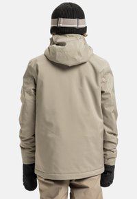 Osoba w beżowej kurtce i spodniach outdoorowych, z czarnymi rękawiczkami i czapką z dzianiny, widziana z tyłu na białym tle.