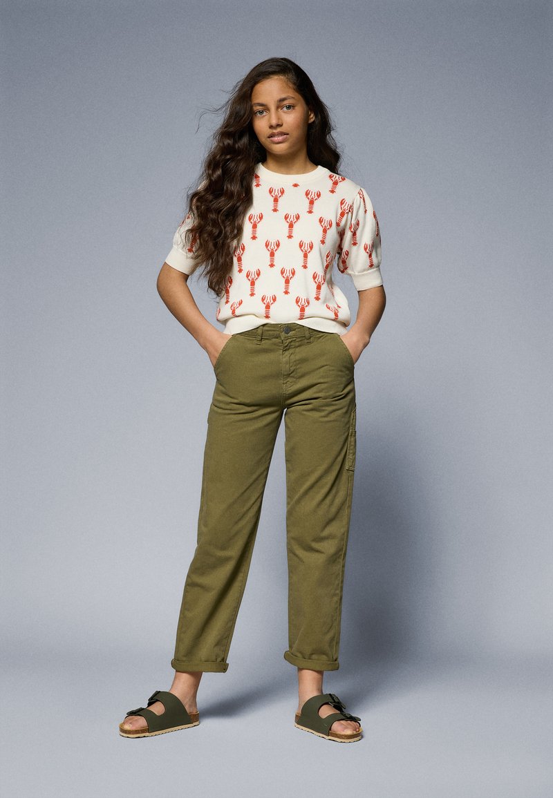 Hellem Pullover mit rotem Hummerdruck, olivgrünen Hosen und dunklen Sandalen. Das Outfit hat eine lockere Passform und umgeschlagene Hosen.