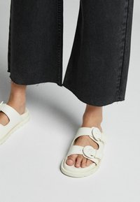 Vita sandaler med två justerbara remmar, rund tå och en texturerad sula, kombinerat med svarta vida jeans med fransad fåll.