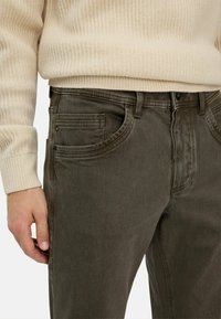 Man in donkere grijze jeans en een crèmekleurige geribbelde trui, met de linkerhand rustend nabij de dij.
