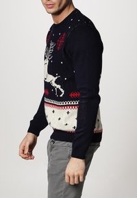 Mörkblå jultröja med renar, snöflingor och geometriska mönster. Tillverkad av stickad tyg, med röda och krämfärgade inslag.