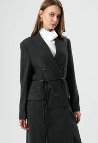 Donkergrijze, op maat gemaakte jas van een wolmix met een visgraatmotief, dubbelgeknoopt ontwerp en een afneembare zwarte ceintuur om de pasvorm aan te passen.