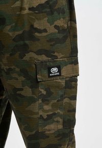 Gros plan sur un pantalon cargo camouflage vert et marron avec une poche à rabat ornée d'un patch logo noir Ecko Unltd.
