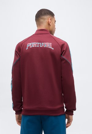 Άνδρας με πλάτη, φοράει μπορντό μπουφάν με την επιγραφή «PORTUGAL» στην πλάτη και μπλε ριγέ σχέδια στα μανίκια.
