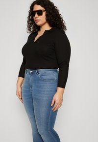 Μαύρο πουκάμισο με μακρύ μανίκι και σχέδιο V, συνδυασμένο με ανοιχτό μπλε τζιν. Το άτομο φορά επίσης μεγάλα ορθογώνια γυαλιά ηλίου.