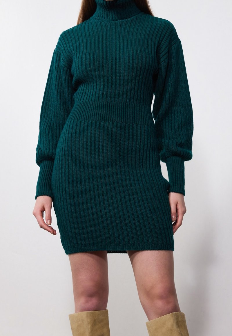 Kvinna i en mörkgrön ribbad klänning med hög hals och långa ärmar, tillsammans med beige mockastövlar, mot en enkel bakgrund.