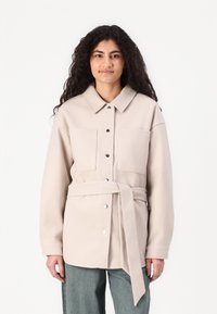Giacca beige chiaro con cintura, due tasche sul petto, bottoni a pressione e colletto a punta; texture morbida e vestibilità rilassata.