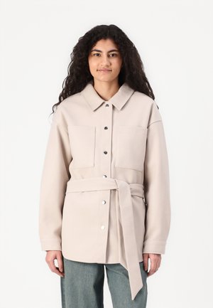 Veste ceinturée beige clair avec deux poches poitrine, boutons-pression et col pointu ; texture douce et coupe décontractée.