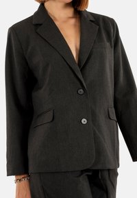 Donna che indossa una giacca di abito a righe scure con due bottoni e tasche a falda, accessoriata con braccialetti colorati di perline.