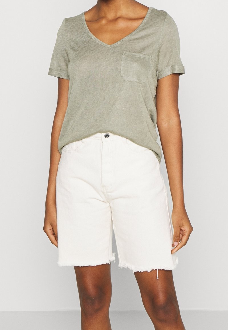 Femme portant un t-shirt vert olive à manches courtes avec col en V et poche poitrine, ainsi qu’un short blanc effiloché à hauteur des genoux.