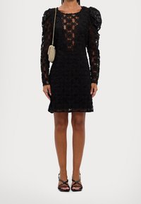 Svart spetsklänning med blommönster, genomskinliga ärmar och en figursydd siluett. Bärs med svarta sandaletter med remmar och en liten, texturerad väska.