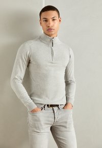 Sweater zippé gris clair avec un col haut, texture en maille fine et poignets côtelés, associé à un jean en denim gris clair.