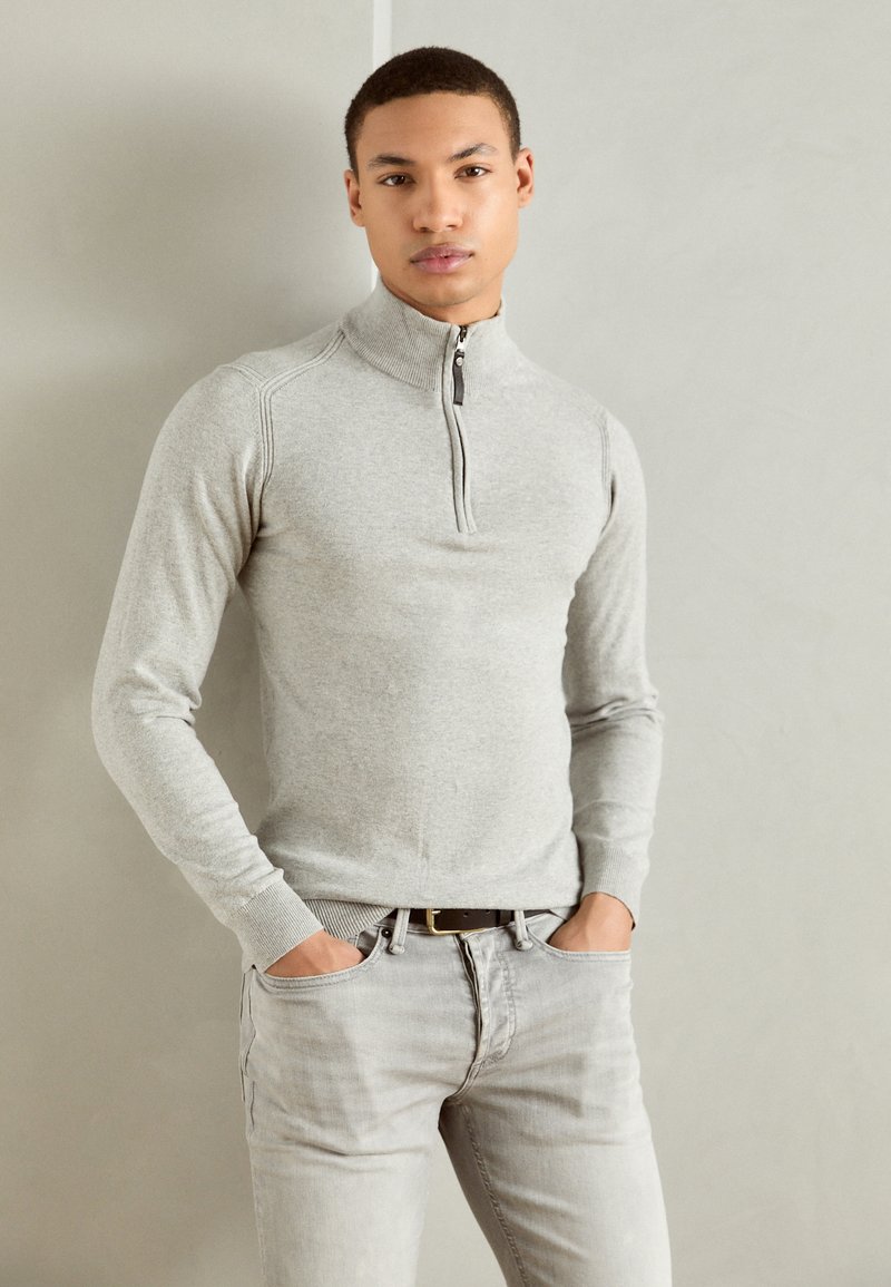 Sweater zippé gris clair avec un col haut, texture en maille fine et poignets côtelés, associé à un jean en denim gris clair.