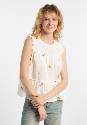 Junge Frau mit lockigem blondem Haar, die eine weiße ärmellose Bluse mit gelben Blumenmustern und hellblaue Jeans trägt und leicht lächelt.