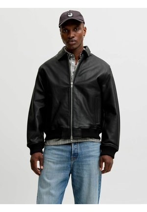 Man met een zwarte leren jas met ritssluiting, grijs geruit overhemd, lichtblauwe spijkerbroek en zwarte pet met logo, staand voor een effen achtergrond.