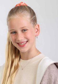 Zilveren ketting met een klein hartje als hanger, lichtbeige trui en een roze scrunchie in blond haar. Glimlachend gezicht met blauwe ogen.