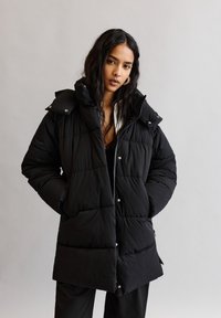 Schwarze Daunenjacke mit hohem Kragen und Druckknopfdetails. Mit gestepptem Design, seitlichen Taschen und einem Reißverschluss.