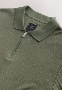 Polo en maille côtelée vert olive avec une petite fermeture éclair et un col doux, avec une étiquette noire portant le nom de la marque NEXT.