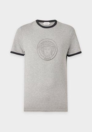 Joven con camiseta gris con ribete negro y logo, pantalones holgados a rayas negras y blancas, y zapatos negros con punteras doradas.
