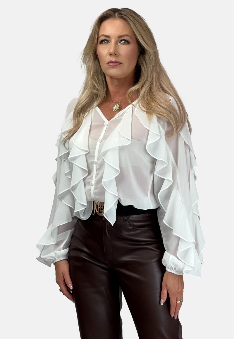 Biała bluzka z szyfonu z falbaniastymi warstwami, długimi rękawami i guzikami. Połączona z brązowymi dopasowanymi spodniami i czarnym paskiem z złotymi akcentami.
