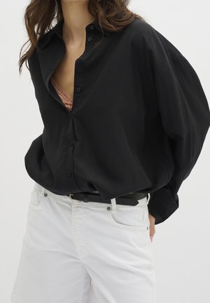 Femme portant une chemise noire ample boutonnée sur un haut en dentelle rose, pantalon blanc avec ceinture noire, mains dans les poches, torse visible.