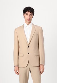 Veste de costume beige en tissu lisse, avec un col cranté, deux poches avant et trois boutons noirs. Chemise blanche en dessous.